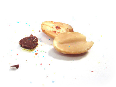 An exploded peanut one. Two nut halves lie naked against each other, some shell fragments are nearby, and a confetti of candy grains litter the floor.